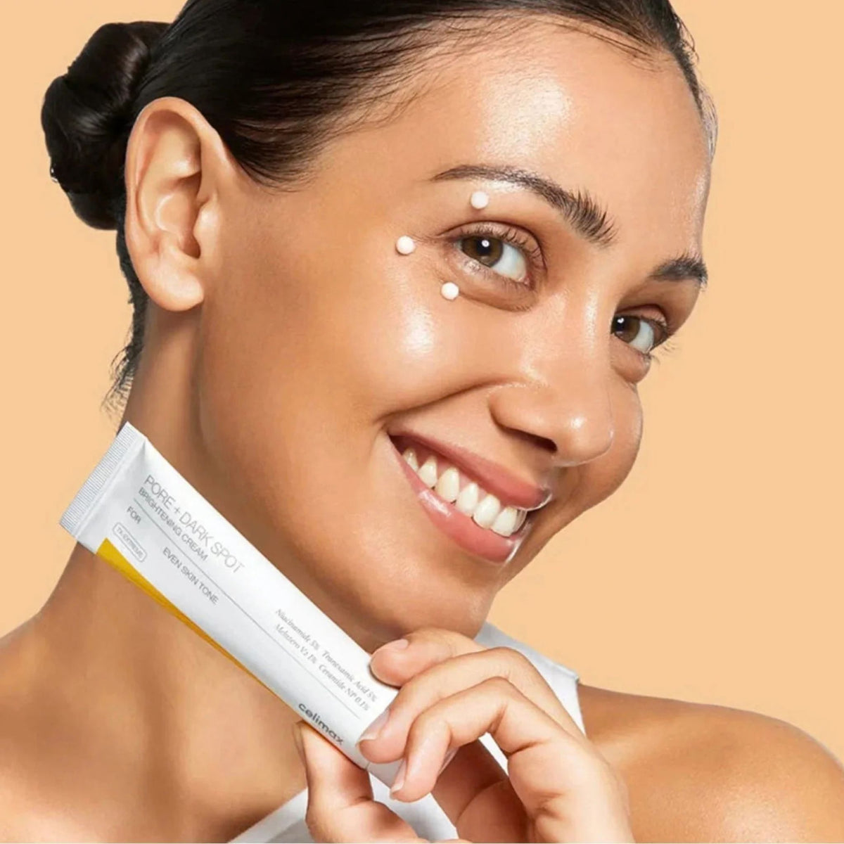 Woman smiling with dots of Celimax Pore + Dark Spot Brightening Cream on her face while holding the tube - K-beauty brightening cream