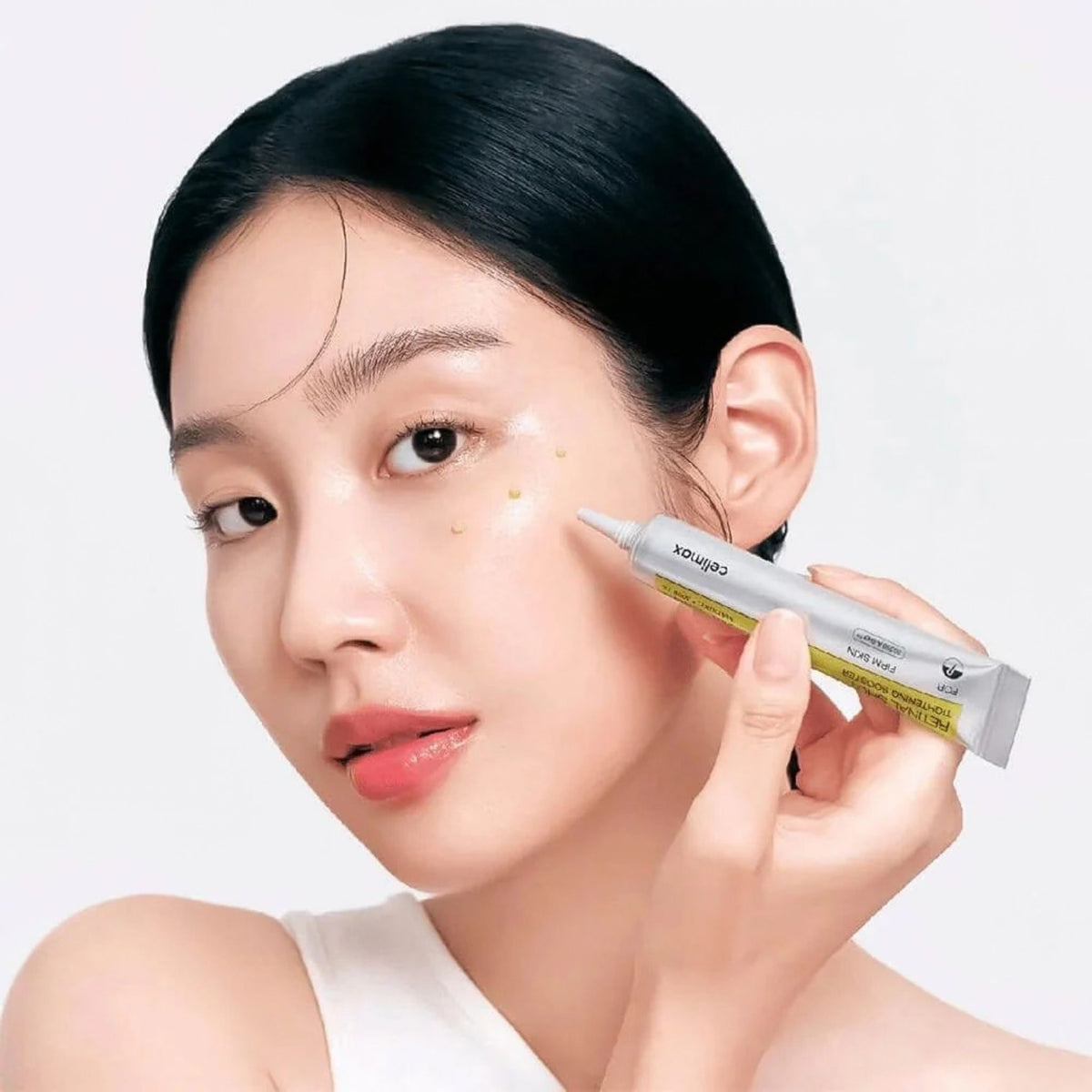 Woman holding Celimax Retinal Shot Tightening Booster tube with small dots of booster on her cheek - K-beauty firming booster