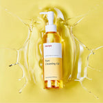 Manyo Pure Cleansing Oil bottle surrounded by oil splashes on yellow background – hydrating cleansing oil for all skin types